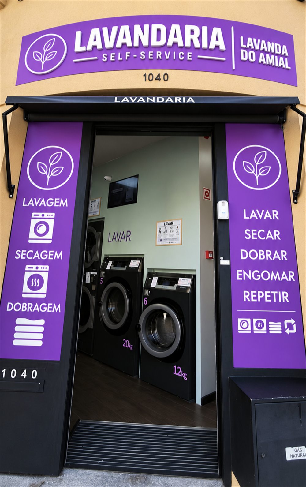 Entrada da Lavanda do Amial: porta aberta com as máquinas de lavar de 20 kg e 12 kg e os painéis roxos Lavagem, Secagem, Dobragem